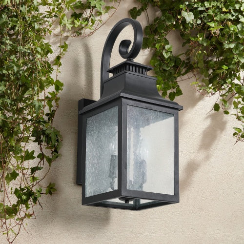 Black outdoor wall lantern with glass panels on a beige wall with green ivy